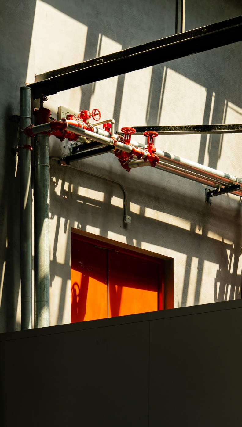Hard sunlight rakes across red fire-protection valves and piping above a red fire door
